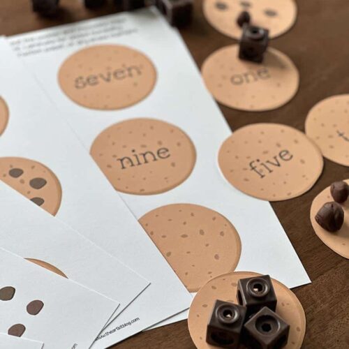 A set of educational counting and number recognition game components with circular cards displaying numbers in words and symbols, wooden cubes, and game tokens on a wooden surface.