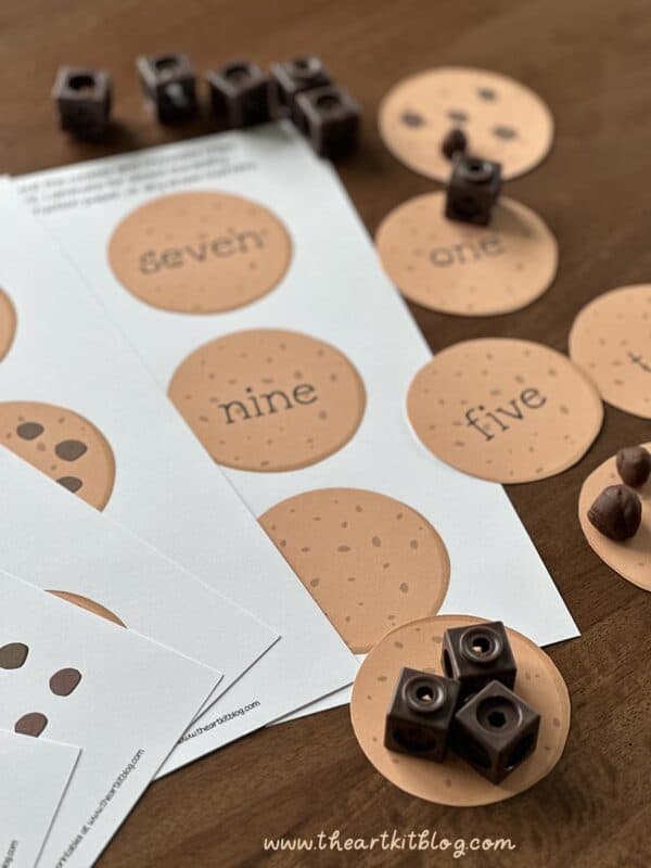cookie-counting-numbers-preschool-math-centers A set of educational counting and number recognition game components with circular cards displaying numbers in words and symbols, wooden cubes, and game tokens on a wooden surface.