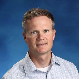 Educator with a friendly expression, wearing a checkered shirt, against a blue background.