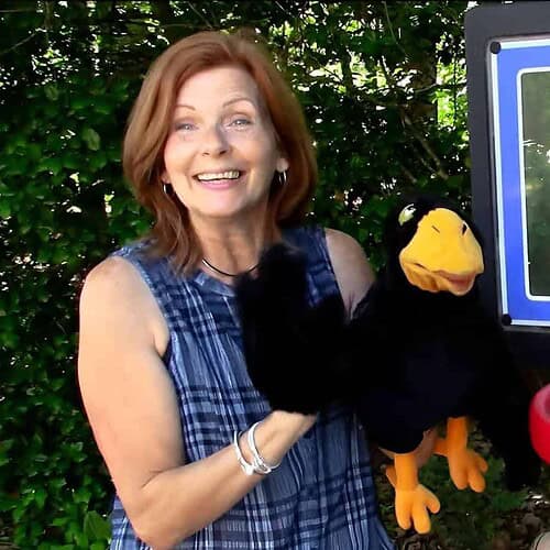 A woman holding a puppet next to a small outdoor basketball hoop and a mini basketball backboard, surrounded by greenery.