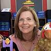 A smiling woman with shoulder-length hair holding a plush turkey puppet in a classroom setting, surrounded by educational materials and colorful storage units.