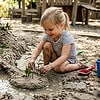 Young girl playing in sandbox, building a small sandcastle with water and toys in an outdoor playground.