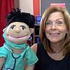 Woman with two puppets, one resembling a nurse and the other a black bird, in a colorful classroom setting.