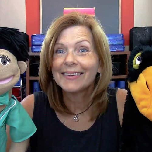 Woman with two puppets, one resembling a nurse and the other a black bird, in a colorful classroom setting.