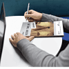cropped-FormIHistoryTimeline_cover2.png Person writing in a homeschool planner with a pen, sitting at a white table.