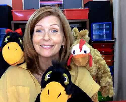 Woman with three colorful plush animal puppets, including a black and yellow bird, a brown bear, and a black and yellow bird, in a classroom setting.