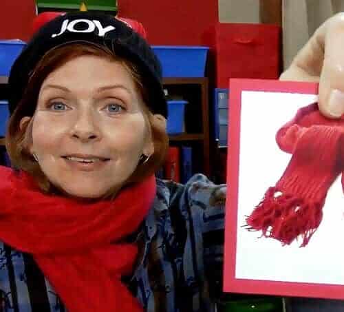 Ms. Denise holding a red knitted scarf with pom-poms, smiling at the camera, in a homeschool setting.