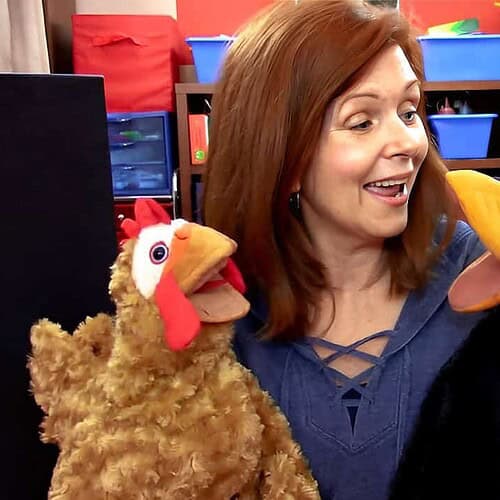 Woman engaging with two puppet chickens in a colorful classroom setting, promoting homeschooling activities.
