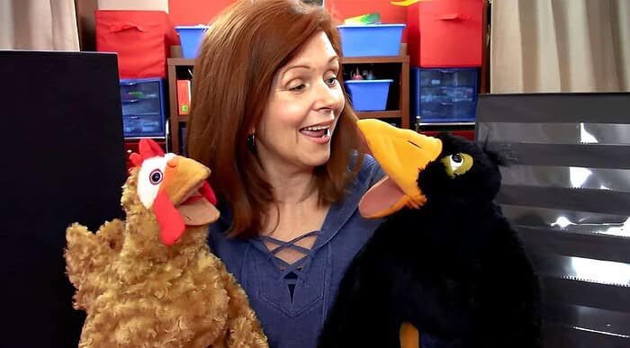 Woman engaging with two puppet chickens in a colorful classroom setting, promoting homeschooling activities.