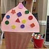 Smiling woman holding a large pink cupcake craft decorated with colorful foam shapes, in a homeschool classroom setting.