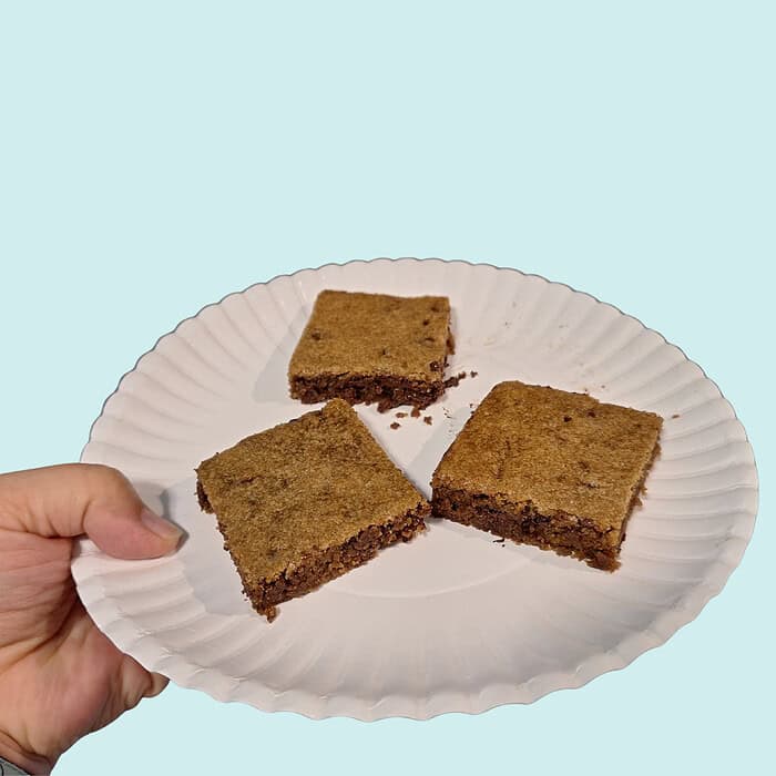blondies bars (2000 x 2000 px) (1) Three homemade brownies on a white paper plate held by a hand against a light blue background.